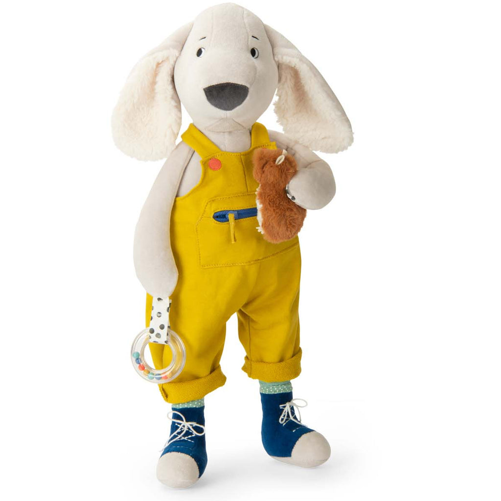 Plush dog toy wearing yellow overalls and blue shoes, holding a clear rattle and fuzzy brown accessory. The image conveys a playful and cheerful tone.