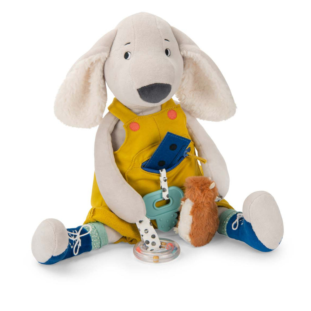 Plush dog toy wearing yellow overalls and blue shoes, holding a clear rattle and fuzzy brown accessory. The image conveys a playful and cheerful tone.