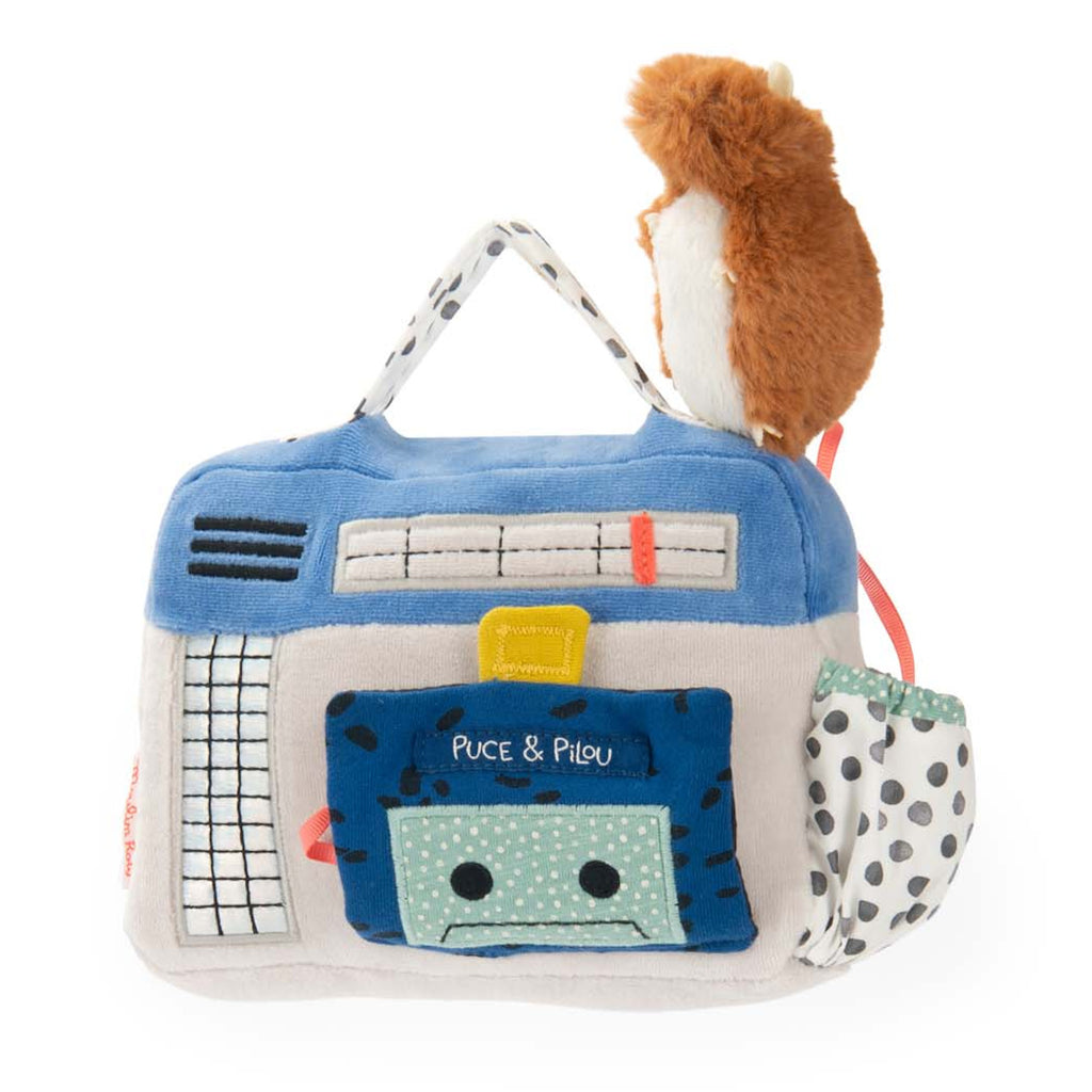 A plush toy resembling a cute animal sits atop a soft, fabric bag designed like a colorful building. The bag features playful patterns and a pocket.