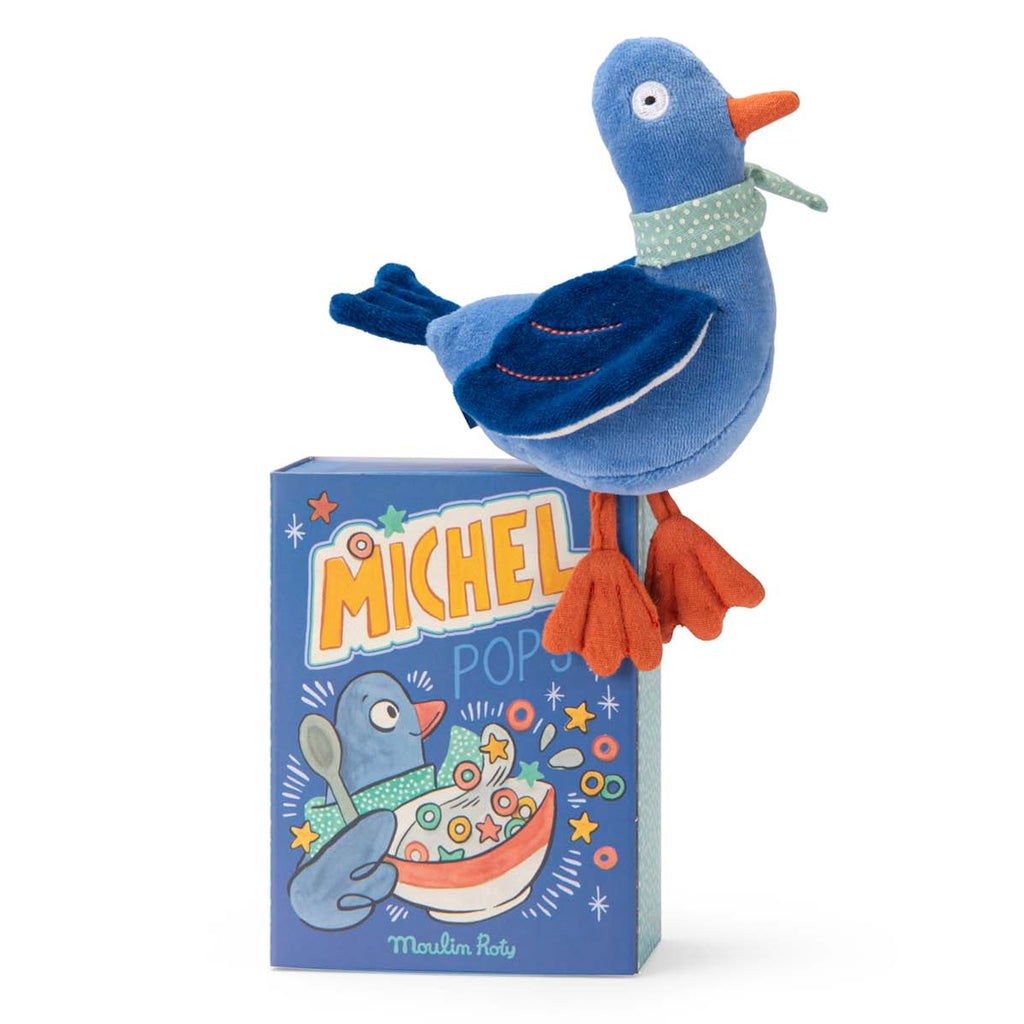 Plush toy blue bird with an orange beak and feet perches on a colorful illustrated box. The scene conveys a playful and whimsical tone.