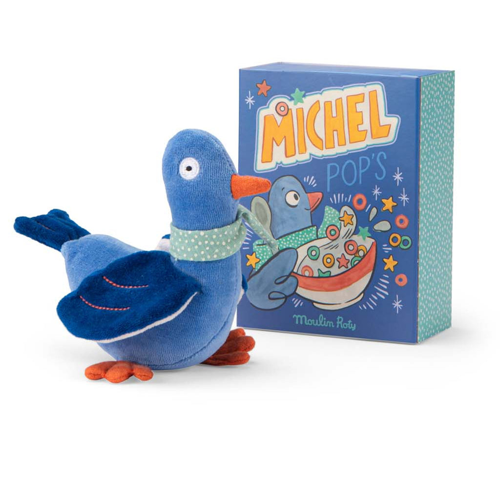 Plush toy blue bird with an orange beak and feet perches on a colorful illustrated box. The scene conveys a playful and whimsical tone.