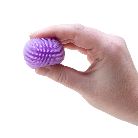 A hand squeezing a small purple sensory ball covered in soft rubber spikes against a white background.