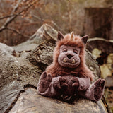 Plush brown horned monster toy sitting outdoors on a large fallen tree log in a forest setting.
