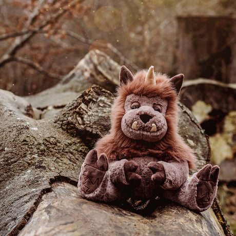 Plush brown horned monster toy sitting outdoors on a large fallen tree log in a forest setting.