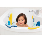 A child with dark hair smiles while playing in a white bathtub filled with water. Colorful bath toys float around, including boats and small figures.