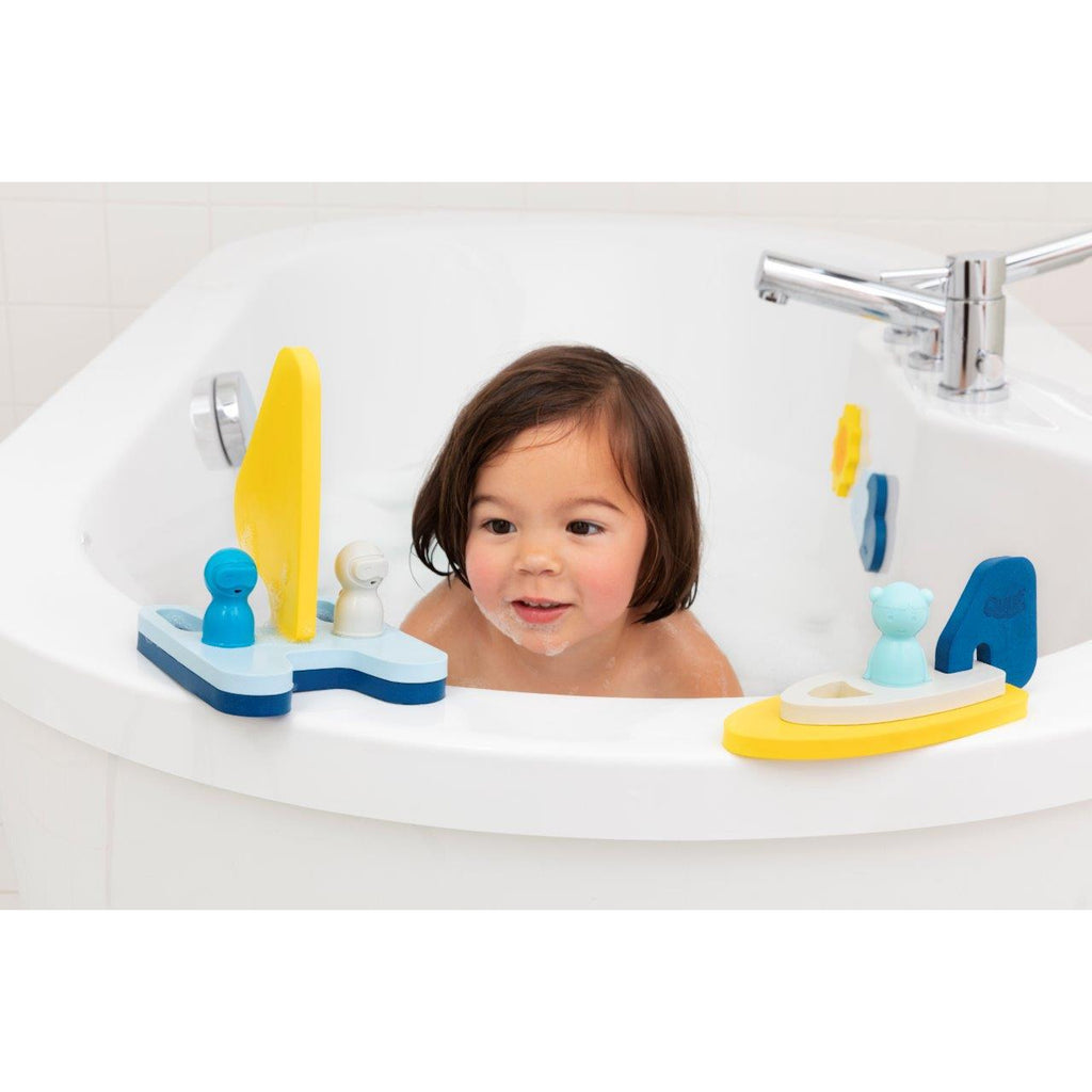 A child with dark hair smiles while playing in a white bathtub filled with water. Colorful bath toys float around, including boats and small figures.