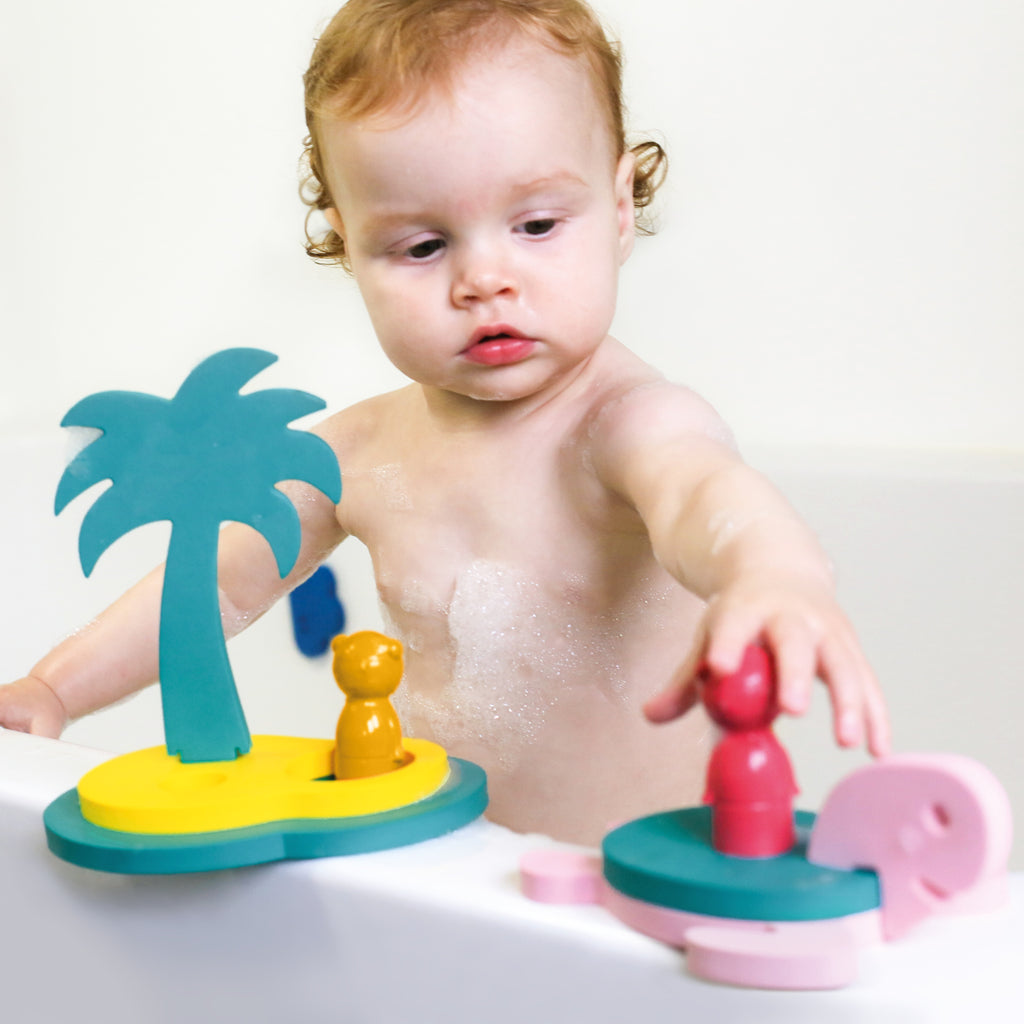 A toddler in a bath plays with colorful foam toys, including a palm tree and animal shapes. The child appears curious and focused.