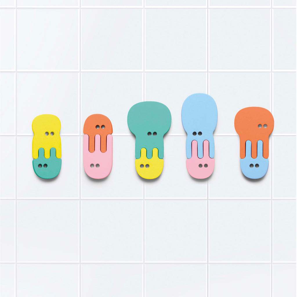 Five colorful, abstract face shapes with button-like eyes hang on a white tiled wall. The shapes are in yellow, pink, green, blue, and orange tones.