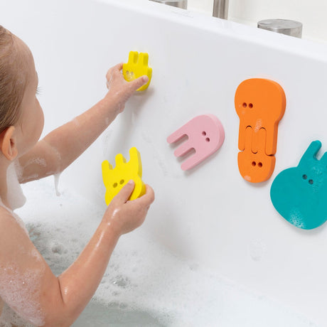 Child in a bubbly bathtub arranging colorful foam jellyfish puzzle pieces on the white tub wall.