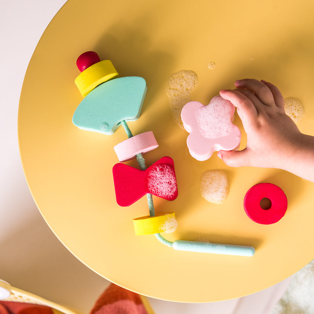 A child's hand plays with colorful foam shapes on a yellow table. The shapes are playful and varied, some with bubbles, creating a fun, cheerful scene.