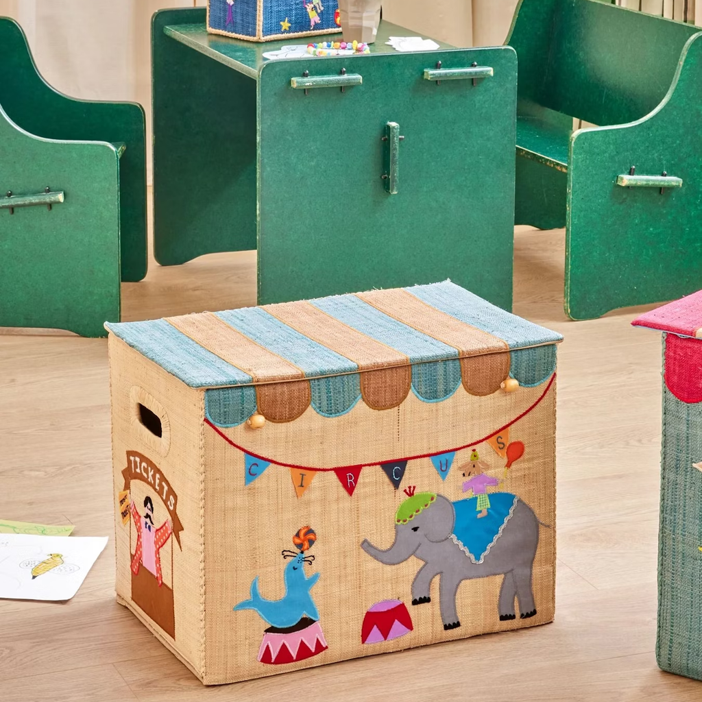 Colorful storage box with circus theme, featuring an elephant and seal illustration. It sits in a room with green chairs and a wooden floor. Fun, playful atmosphere.