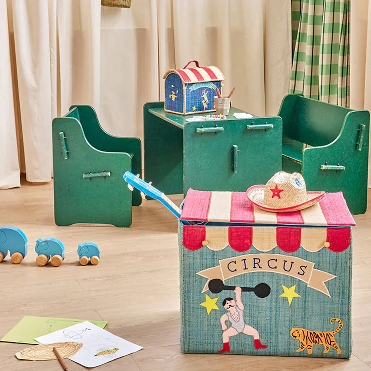 Children's playroom with circus-themed decor. A colorful toy chest, green chairs and table, toy elephants, and drawings create a whimsical, playful atmosphere.