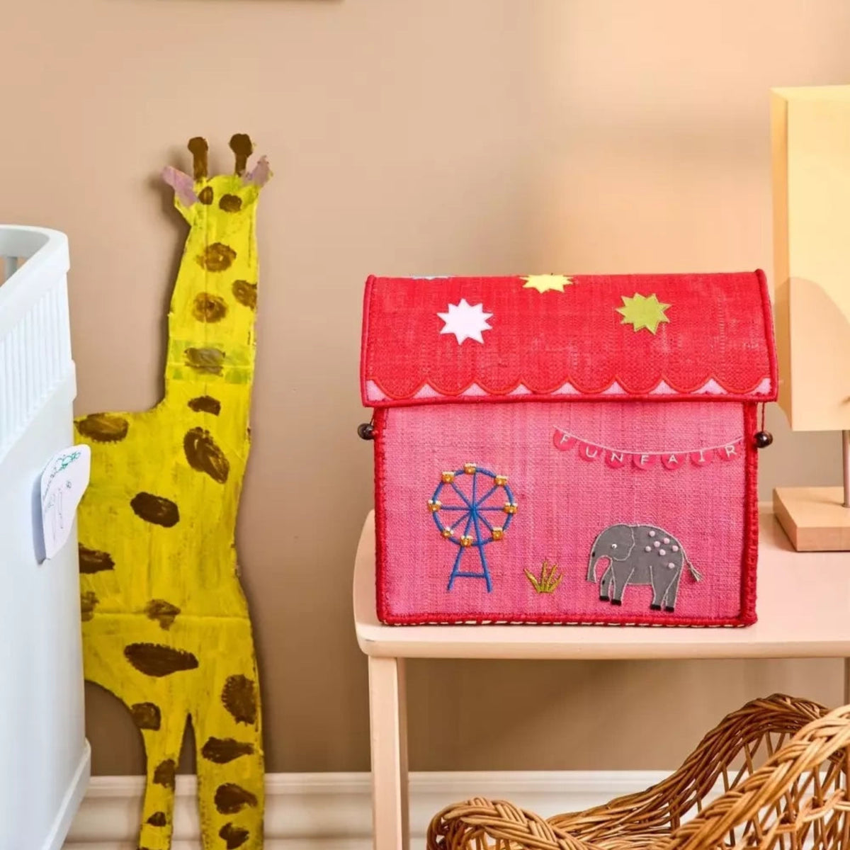 Red and pink raffia toy basket shaped like a house with funfair decorations on a table in a nursery.