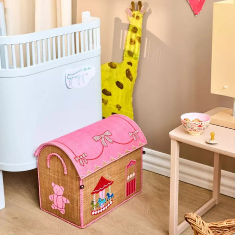 A pink-lidded raffia toy basket shaped like a house sits on the floor of a child's nursery room.