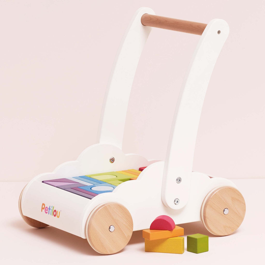 Wooden baby walker with a white frame, natural wood wheels, and rainbow-colored blocks. The design is playful and cute, set against a light pink background.