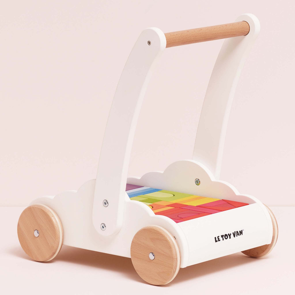 Wooden baby walker with a white frame, natural wood wheels, and rainbow-colored blocks. The design is playful and cute, set against a light pink background.