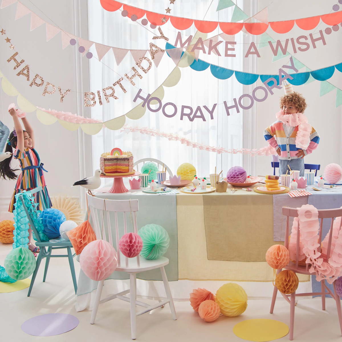Children celebrating at a party table decorated with pastel honeycomb balls, garlands, and a birthday cake.