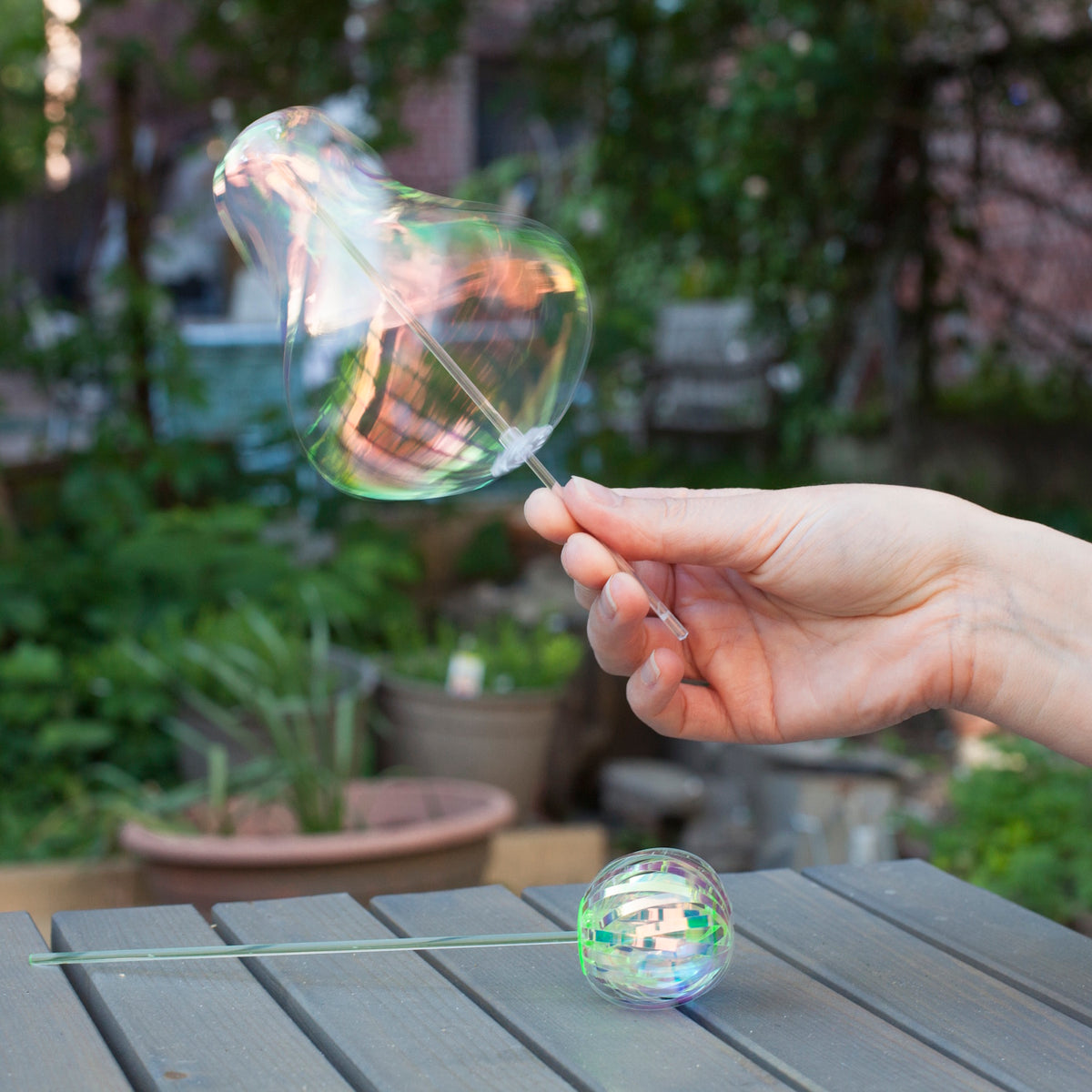 A hand holds a wand with a shimmering, translucent bubble in a garden setting. A round container rests on a wooden table, evoking a playful, serene mood.