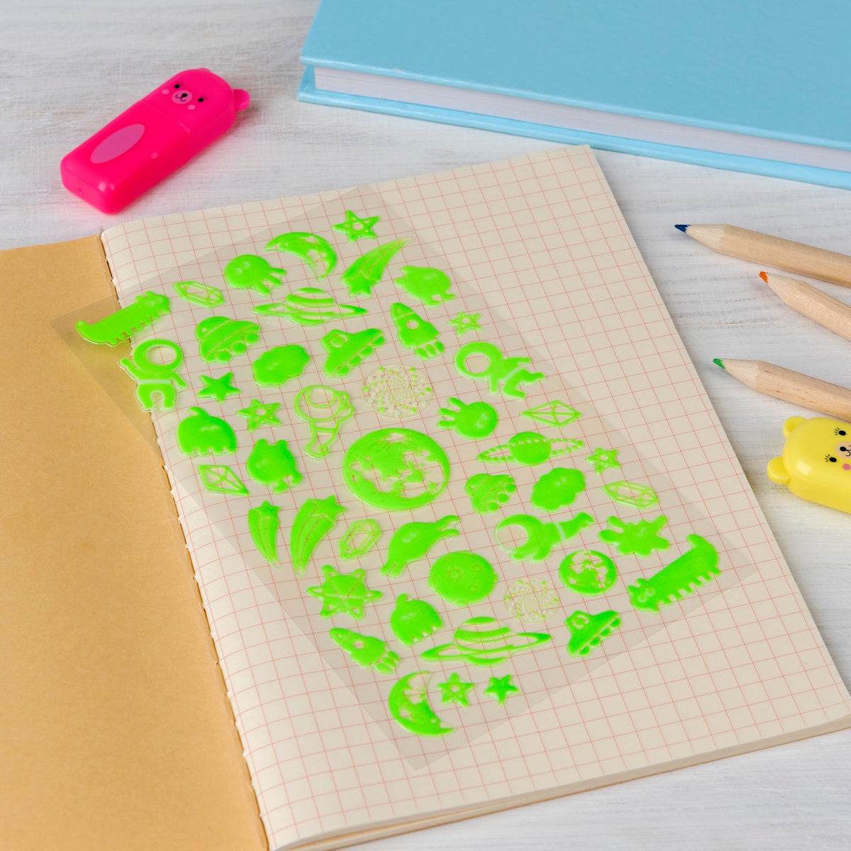 Notebook with a grid pattern open on a table, displaying neon green space-themed stencils. Nearby are colored pencils and a pink eraser, evoking creativity.