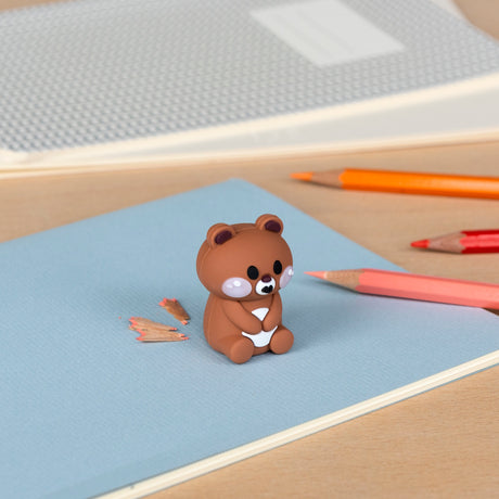 A cute bear-shaped eraser sits on a blue notebook with scattered pencil shavings. Nearby, several colorful pencils create a playful, creative vibe.