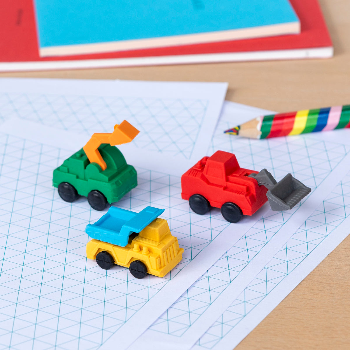 Toy vehicles in primary colors on graph paper. A green excavator, red bulldozer, and yellow dump truck. Nearby are a colorful pencil and red notebook. Playful tone.