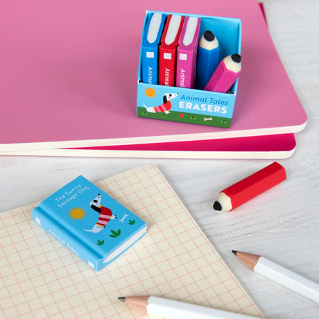A playful stationery set with colorful erasers shaped as books and pencils is displayed on a table. Two pink notebooks add a cheerful touch.