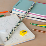 A playful scene on a wooden desk features an open notebook with tree sketches, a decorative pencil, colorful sharpeners, and stacked pastel notebooks.
