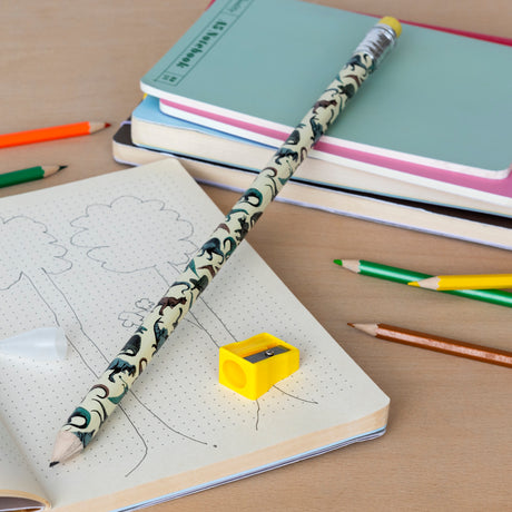 A playful scene on a wooden desk features an open notebook with tree sketches, a decorative pencil, colorful sharpeners, and stacked pastel notebooks.
