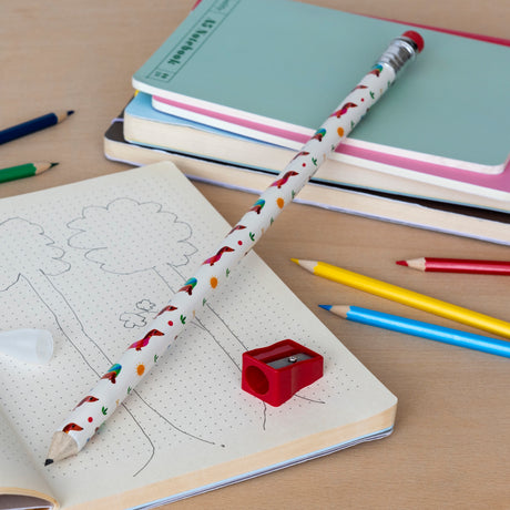 A whimsical scene with a large, printed pencil and red sharpener on a dotted notebook featuring tree sketches. Colored pencils and pastel notebooks add a playful touch.