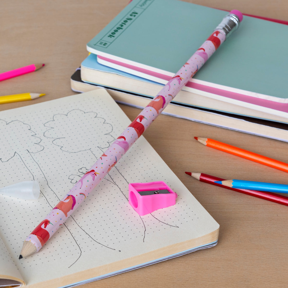 A pink, patterned oversized pencil on a notebook with tree sketches. Bright colored pencils, sharpener, and notebooks surround it on a wooden surface.