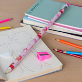 A pink, patterned oversized pencil on a notebook with tree sketches. Bright colored pencils, sharpener, and notebooks surround it on a wooden surface.