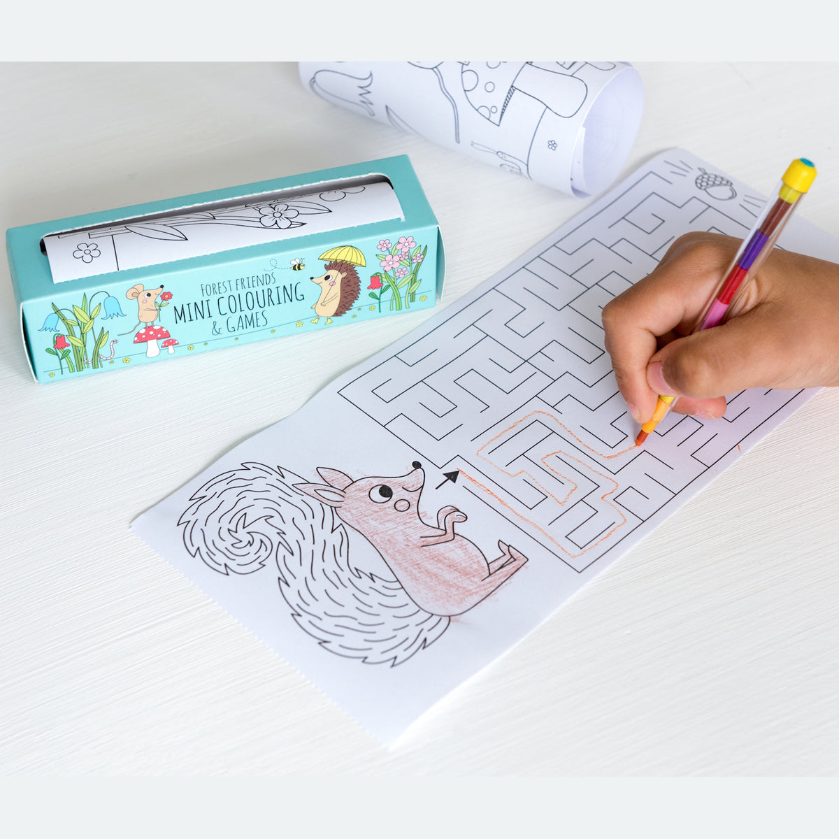 Child's hand using a multicolored pencil to complete a maze on a woodland-themed coloring sheet featuring a squirrel. A package labeled "Forest Friends Mini Colouring & Games" is nearby.