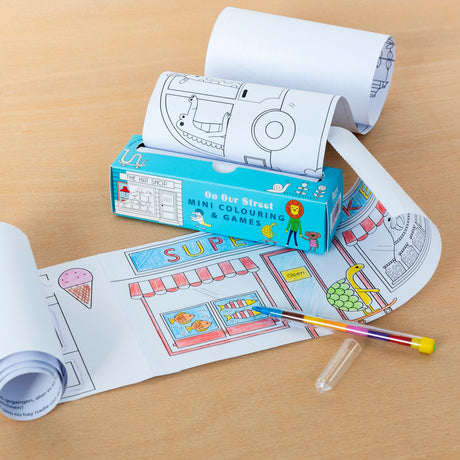 A colorful, playful image of a children's mini coloring and games set named "On Our Street." Rolled coloring sheets feature a street scene with a shop, partially colored with blue and red. A nearby pen adds to the creative, joyful atmosphere.