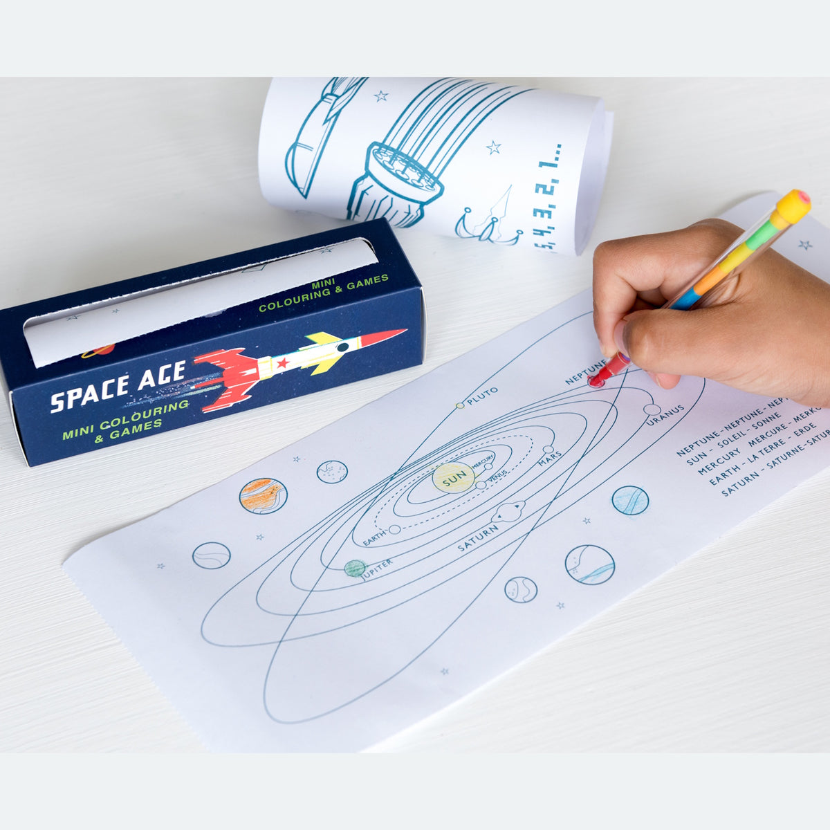 A child's hand colors a solar system worksheet with a multicolored pencil. Nearby, a “Space Ace” mini coloring set with rocket artwork lies open, evoking creativity.