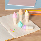 Five ice cream cone-shaped highlighters in pastel colors are on a checkered notebook. Two pencils and closed notebooks are in the background.