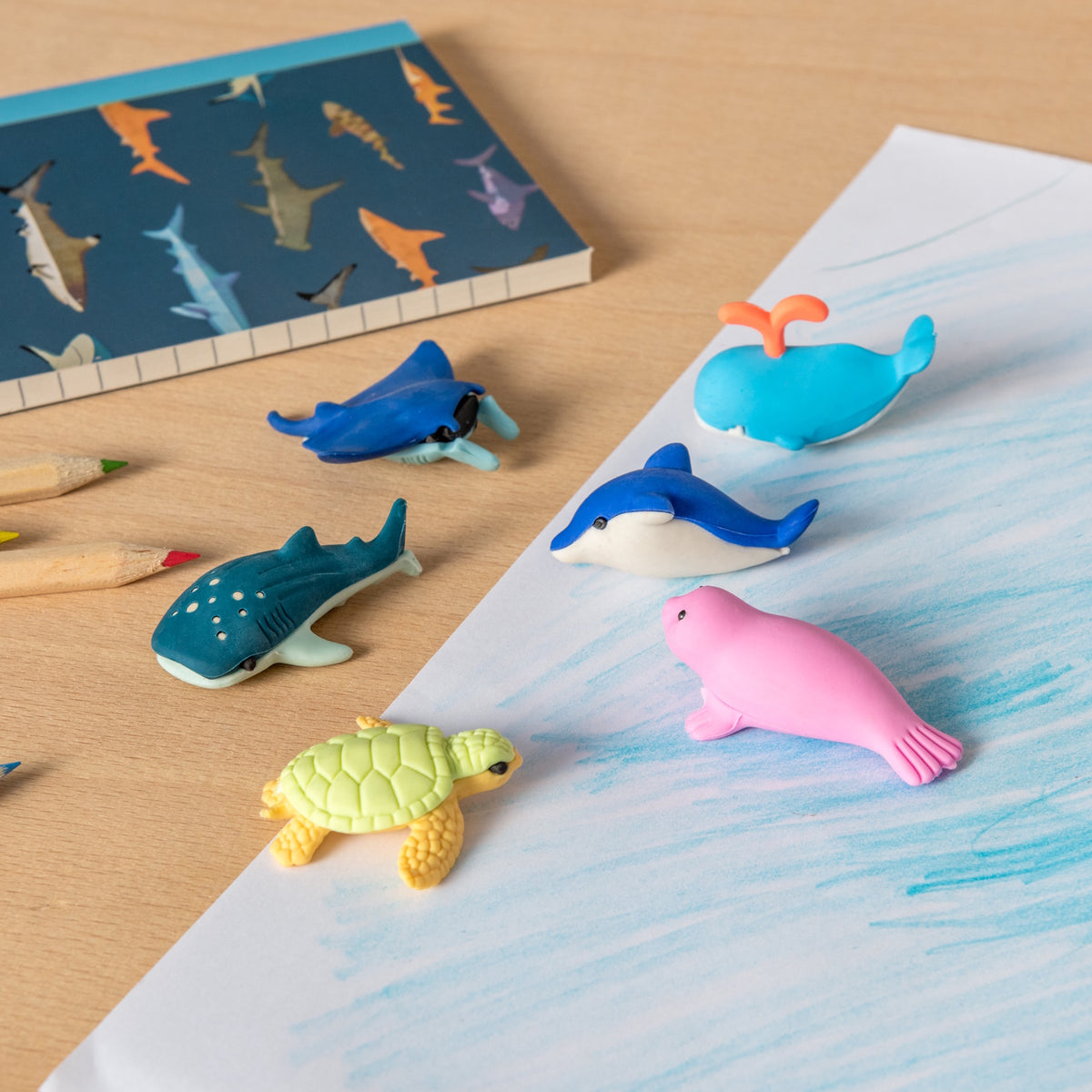 Toy sea animals, including a turtle, whale, dolphin, and seal, are arranged on a desk with colored pencils and a shark-patterned notebook. Playful vibe.