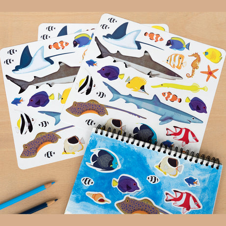 Sticker sheets featuring vibrant marine life, like sharks, rays, seahorses, and colorful fish, lie beside a sketchbook with similar ocean stickers. Two blue pencils complement the scene.