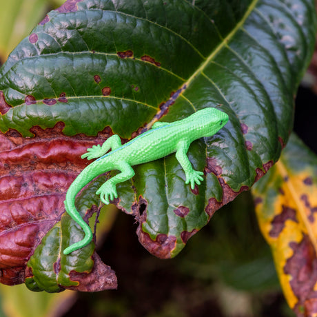A bright green lizard toy lies on a large, dark green leaf with brown patches. The scene has a vibrant, natural feel with rich textures.