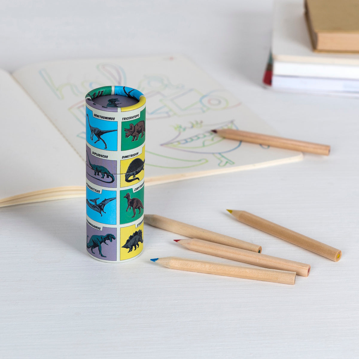 A colorful pencil holder with dinosaur illustrations stands on a table amid scattered colored pencils. An open coloring book shows a whimsical design.