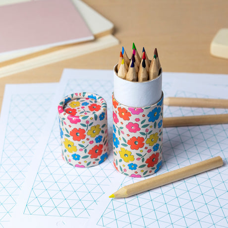 A floral-patterned cylindrical case holds colorful pencils on a desk with geometric grid paper, conveying a creative and organized workspace vibe.