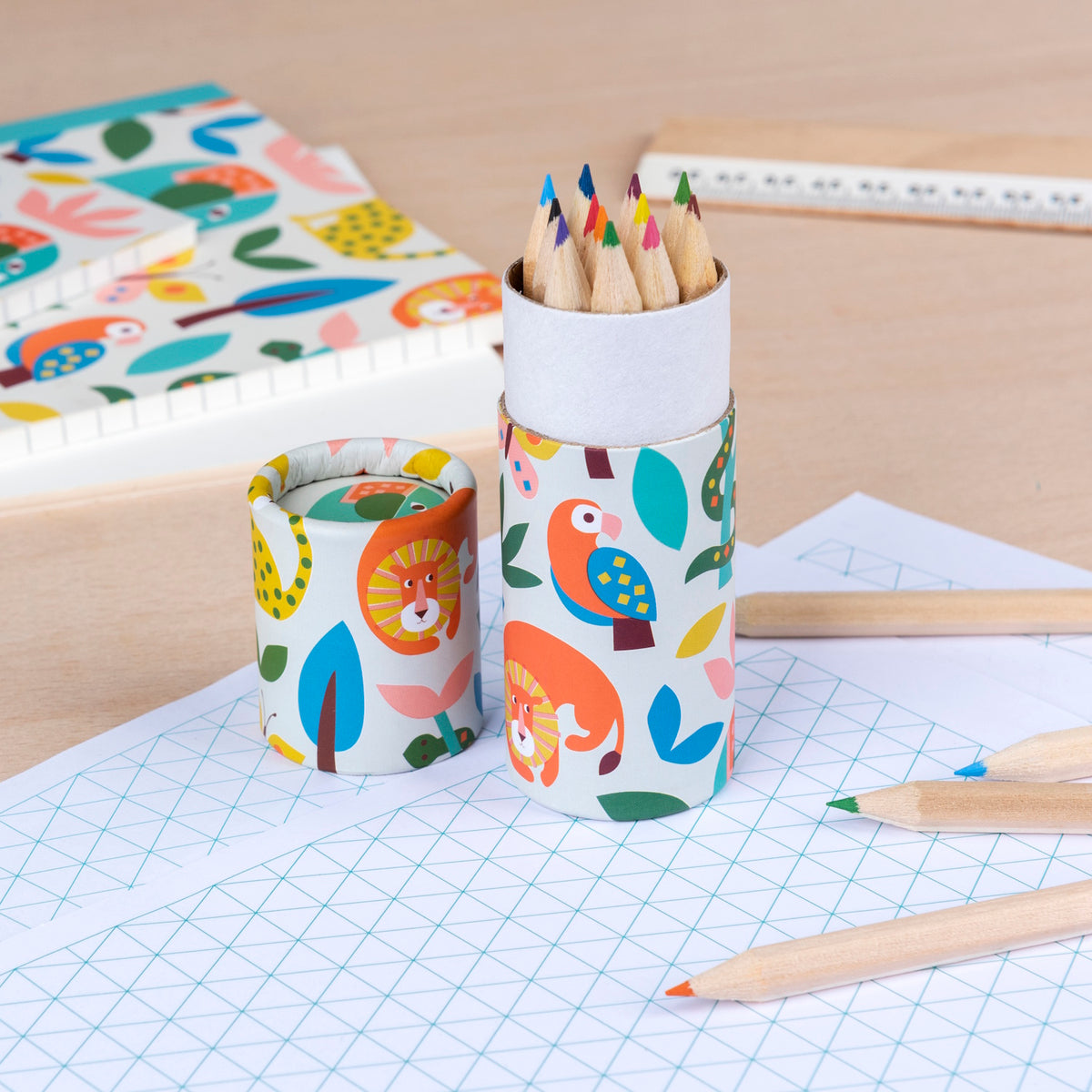 A set of colorful pencils in a tube with vibrant animal graphics sits on a wooden desk with grid paper and a ruler, evoking a playful, creative mood.