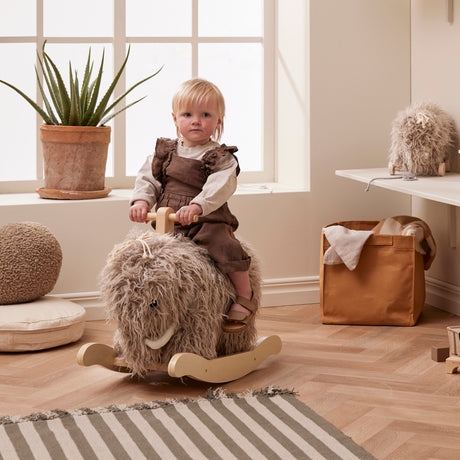 Toddler on a plush mammoth rocking horse toy in a neutral nursery room.