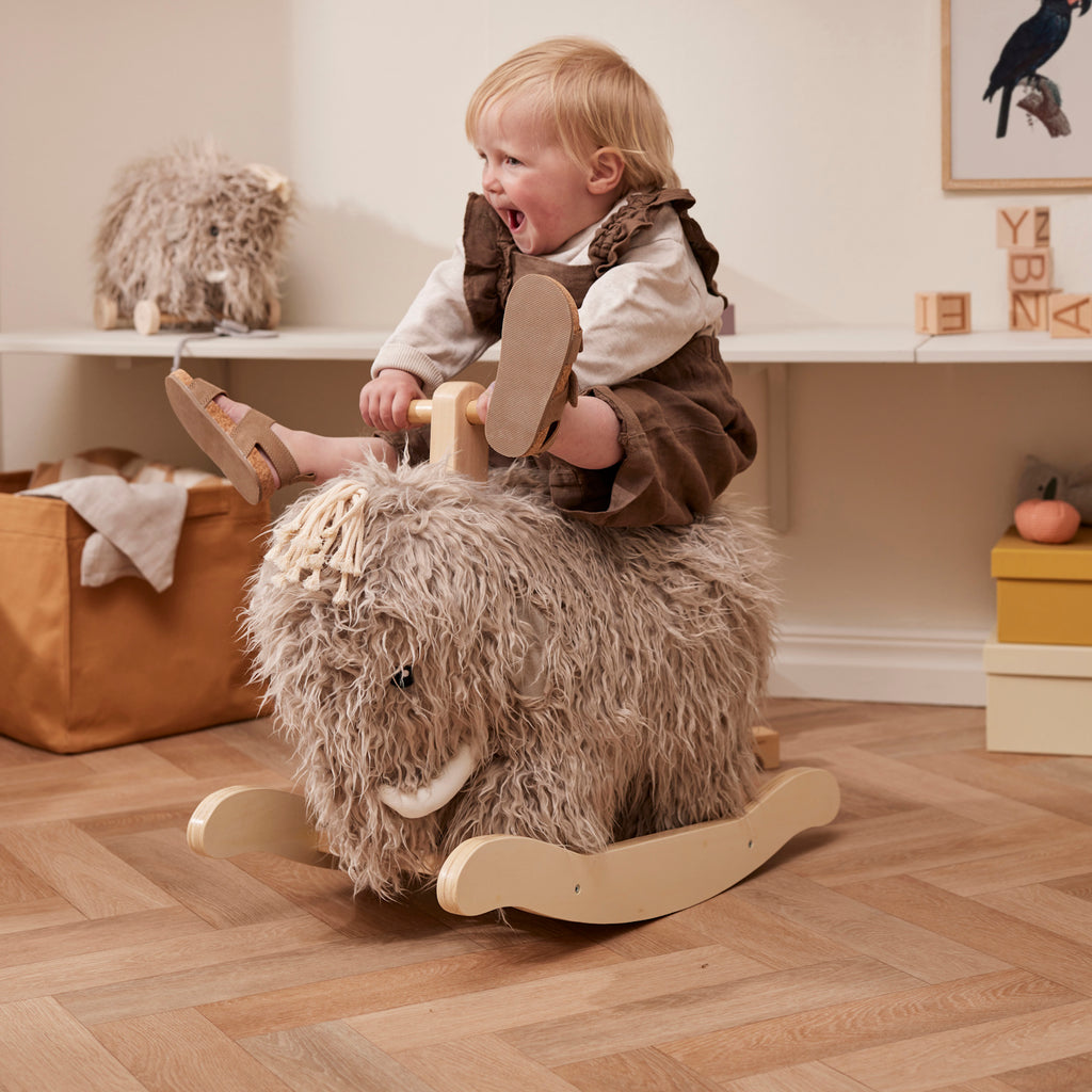 A joyful toddler rides a furry mammoth-shaped rocking toy in a cozy room. The child is laughing, exuding happiness and playfulness.