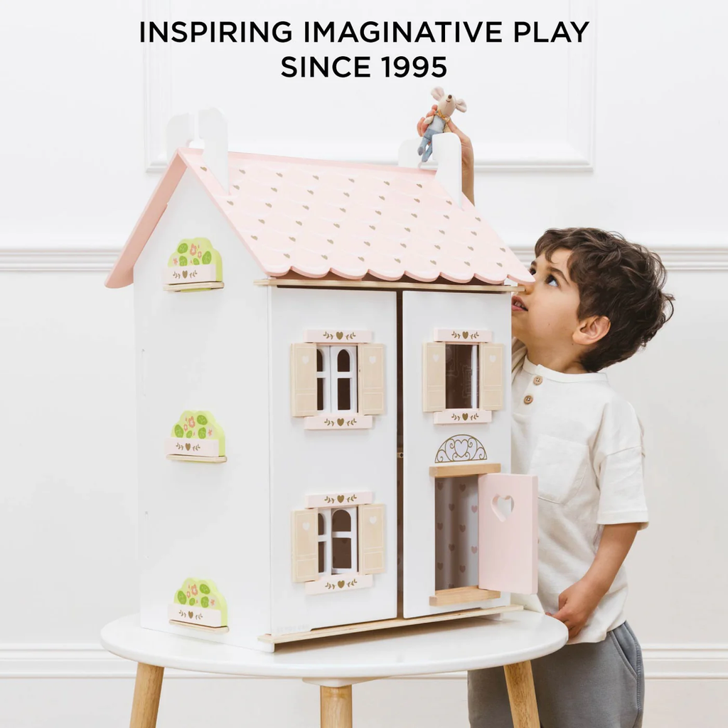 A young child in a white shirt plays with a large, pastel-colored dollhouse on a table. The text above reads, "Inspiring Imaginative Play Since 1995."