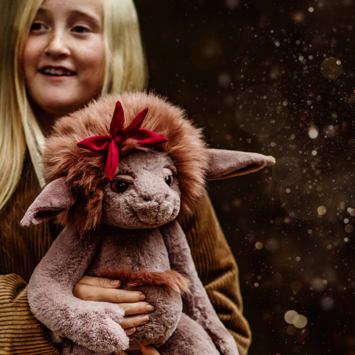 Smiling blonde girl holding a plush creature with a red bow, brown mane, and floppy ears in a magical setting.