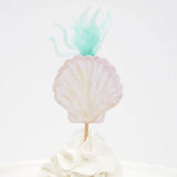 A seashell-shaped topper with green seaweed details on top of a frosted white cupcake.