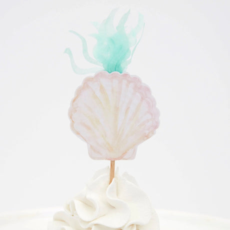 A seashell-shaped topper with green seaweed details on top of a frosted white cupcake.