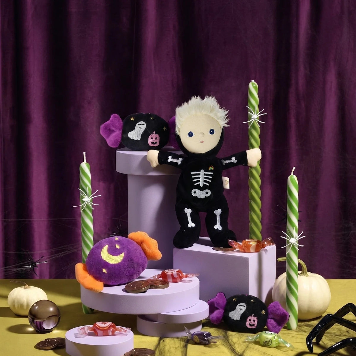 Skeleton plush doll displayed with Halloween decor, candies, and candles against purple curtains.
