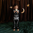 Young child in a skeleton outfit holding a matching skeleton plush doll against dark curtains.