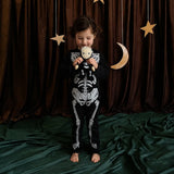 Young child in a skeleton outfit holding a matching skeleton plush doll against dark curtains.